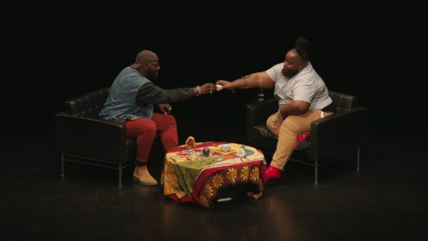 Two people sit on a stage across from each other with a coffee table between them; they cheers with two small cups.