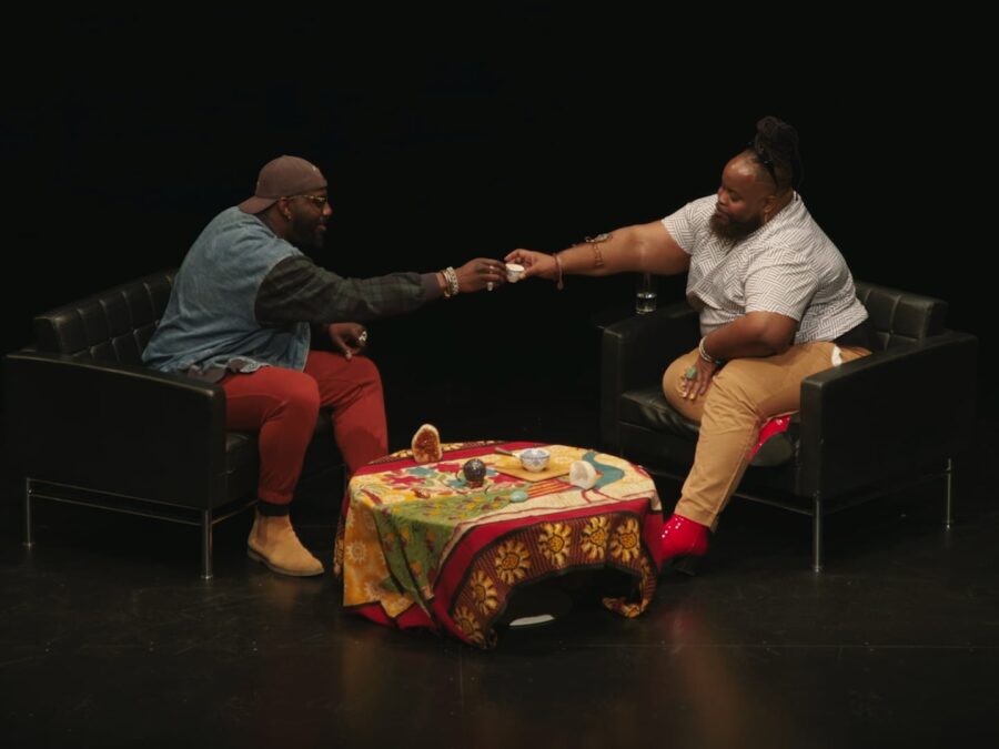 Two people sit on a stage across from each other with a coffee table between them; they cheers with two small cups.