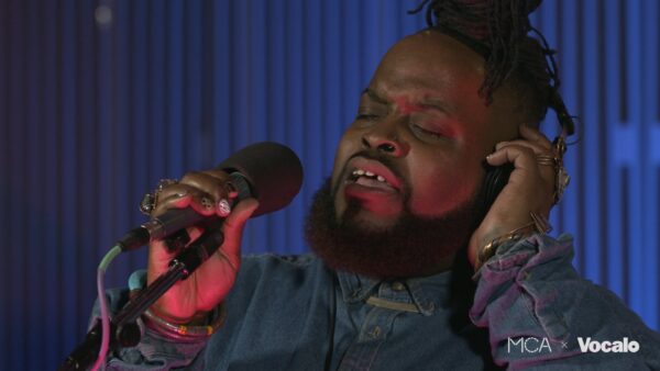 Close up of a dark skinned person with a beard singing into a microphone as they cover their left ear. In the corner, small white text reads Vocalo.