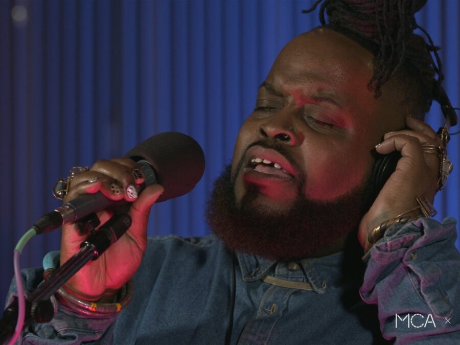 Close up of a dark skinned person with a beard singing into a microphone as they cover their left ear. In the corner, small white text reads Vocalo.