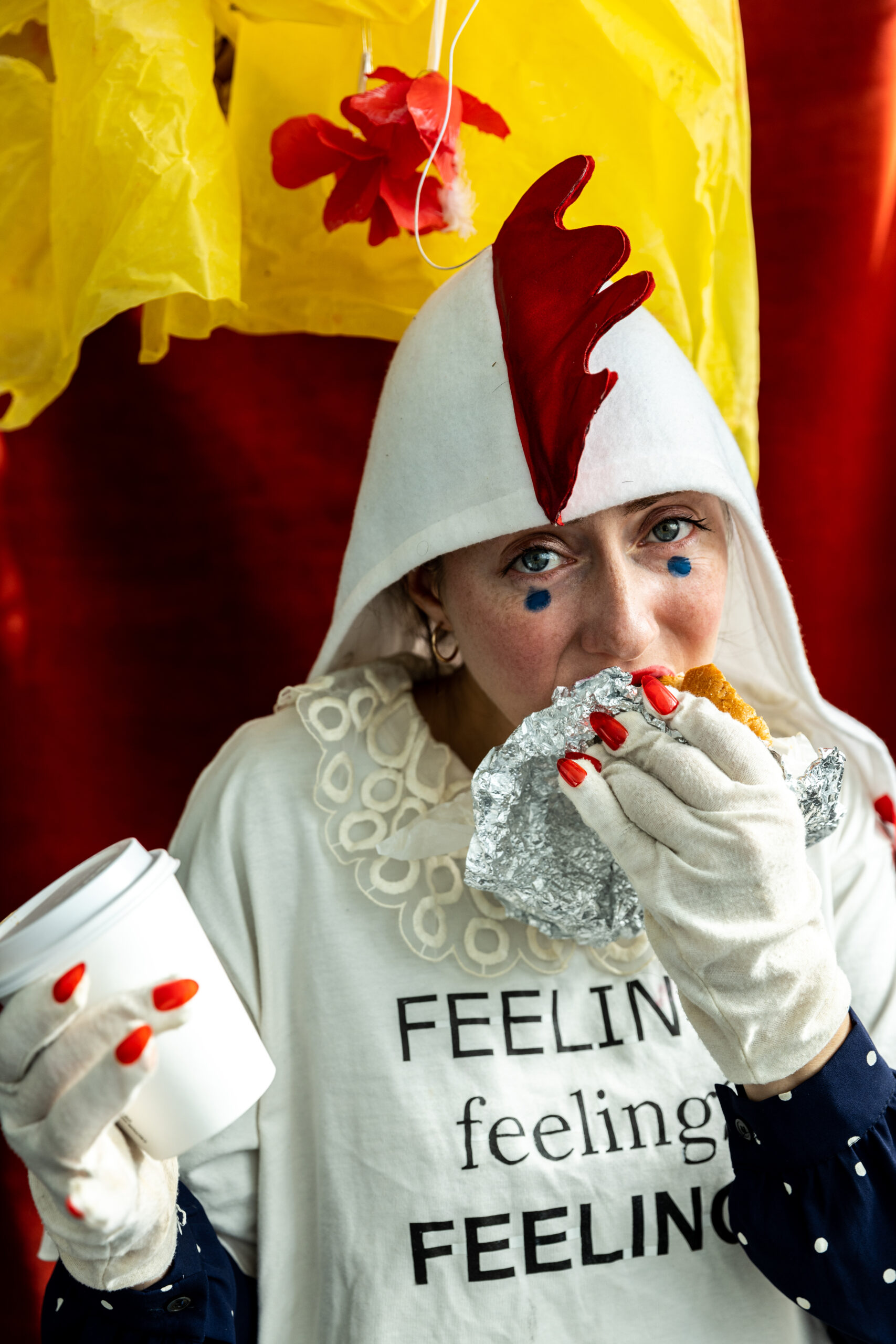 A person in a chicken costume eats a sandwich wrapped in tin foil while holding a coffee and wearing a white t-shirt that partially reads Feelin, feeling, feeling