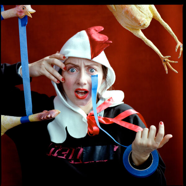 A person in chicken hood looks at the camera in bewilderment as rubber chickens seemingly float in the air around them, some connected by blue painters tape