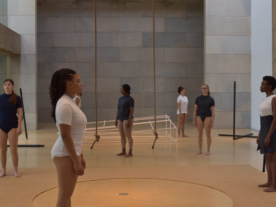 Six individuals stand in a large, minimalist room with concrete walls and a light-colored floor. Most wear form-fitting tops and bottoms, while one wears a white top and dark skirt. Two ropes hang from the ceiling, and a white rectangular frame, two black poles, and a circular mat are on the floor.