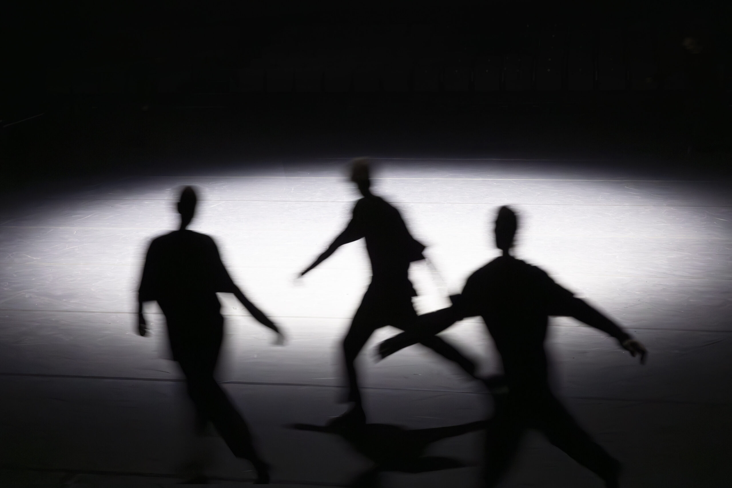 Three silhouetted figures are moving around in a dimly lit setting, highlighted only by a bright oval light.