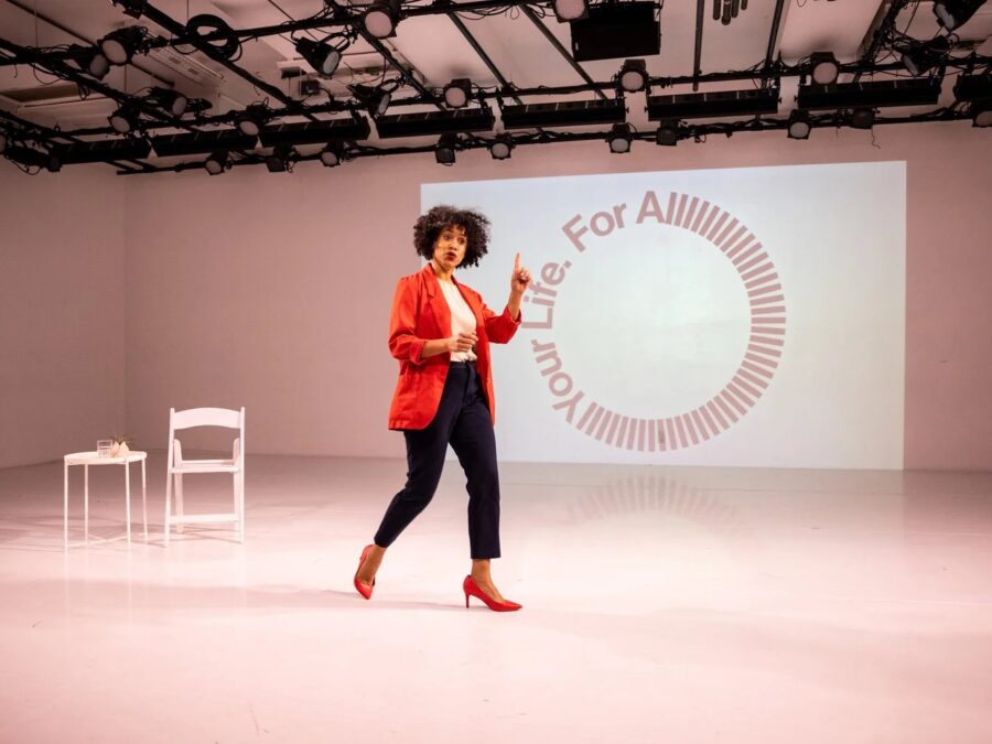 A presenter dressed in a red blazer, black cropped pants, and red shoes raises one finger as if making a point in front of a slide projected on a white wall with text displayed in a circle that reads For All Your Life. The word all has many l's in it, to drag the word on.
