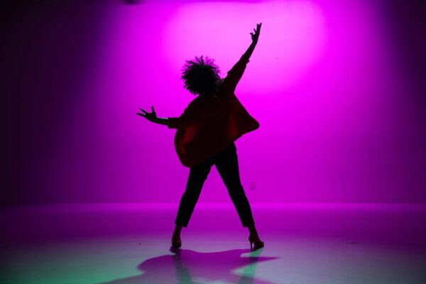 The silhouette of a person with a curly afro, posed with arms and legs spread in a showcase style against a magenta-lit background