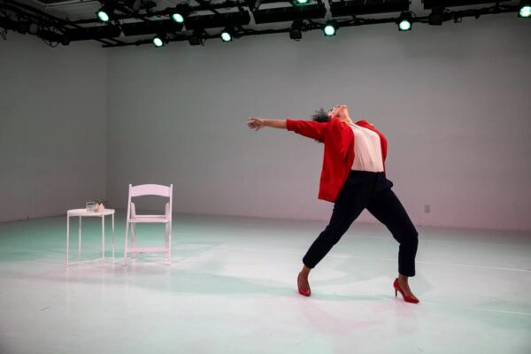A person in a red blazer, white shirt, and navy trousers posed in a dramatic motion, head tilted backward as they point backward toward a white chair and a small table nearby on the stage. Green stage lights are visible in the background.