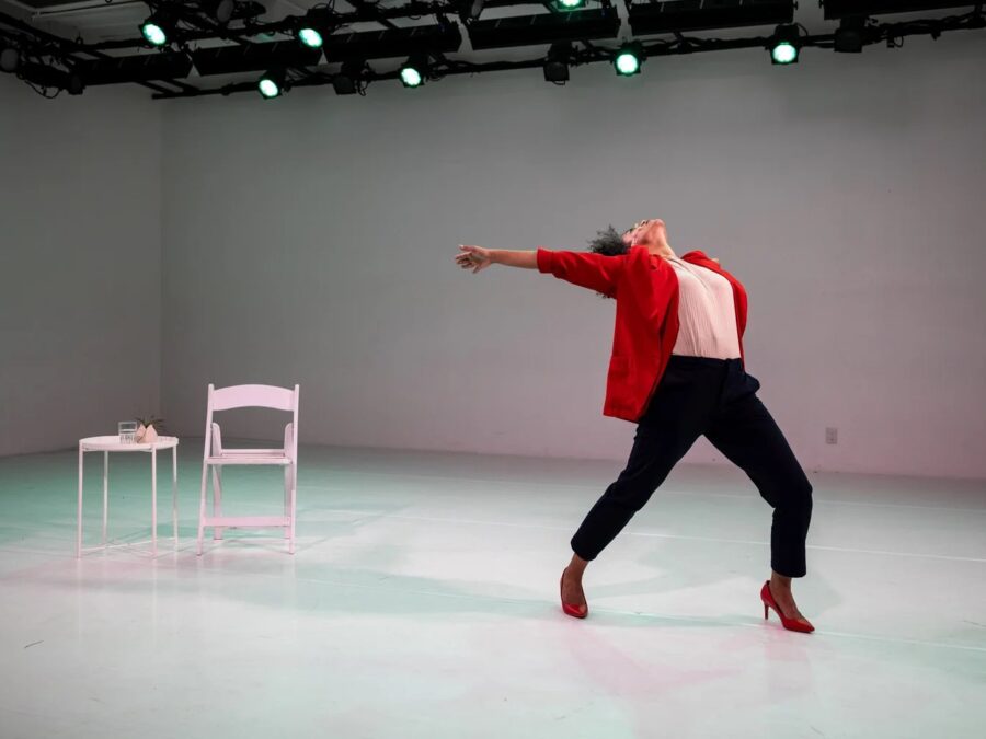 A person in a red blazer, white shirt, and navy trousers posed in a dramatic motion, head tilted backward as they point backward toward a white chair and a small table nearby on the stage. Green stage lights are visible in the background.