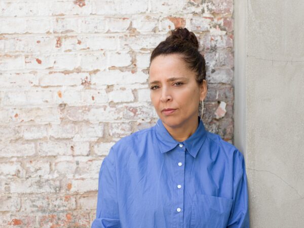A female-presenting person in a blue collared shirt with their brown hair pulled into a bun poses against a white-washed brick wall
