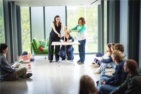 Three people engage with a humanoid puppet on a table in a room with an audience sitting on the floor