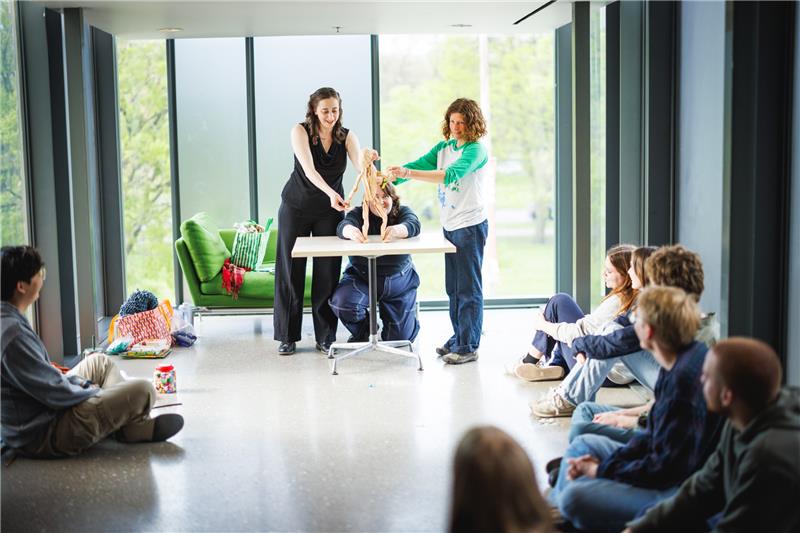 Three people engage with a humanoid puppet on a table in a room with an audience sitting on the floor