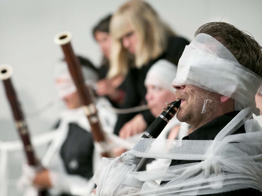 Musicians playing wooden recorders, seated in a row, wrapped in white gauze, some around their head as well.