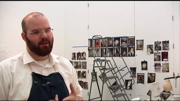 A male presenting person in an apron stands in front of a step ladder with several pictures hung on a white wall