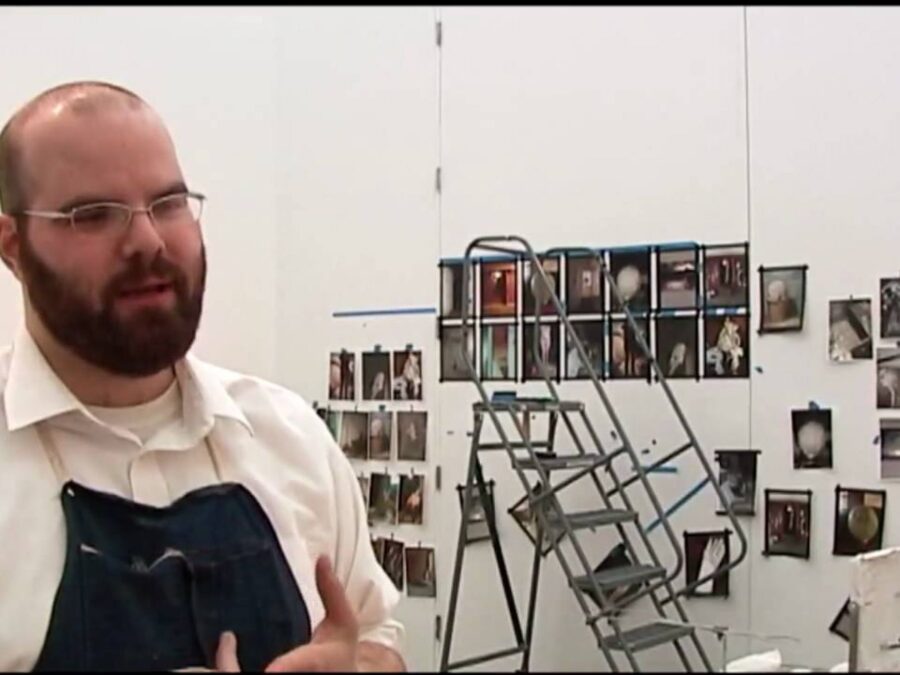 A male presenting person in an apron stands in front of a step ladder with several pictures hung on a white wall
