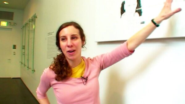 A female presenting person with brown curly hair pulled back in a ponytail points to an artwork hanging on a wall with her left hand