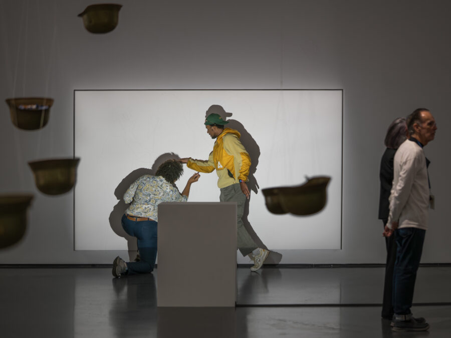 A person traces another person's shadow posed in a walking motion on a white wall in a room filled with hanging helmets