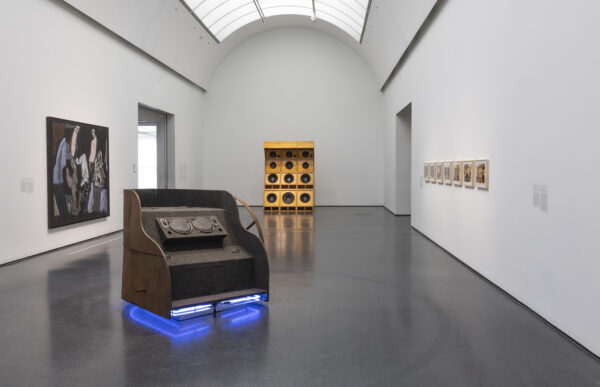 A museum gallery features a dark, upholstered sculpture with embedded speakers and a glowing blue base. Behind it stands a tall, wooden stack of speakers, while a large painting and a row of framed artworks hang on the white walls.