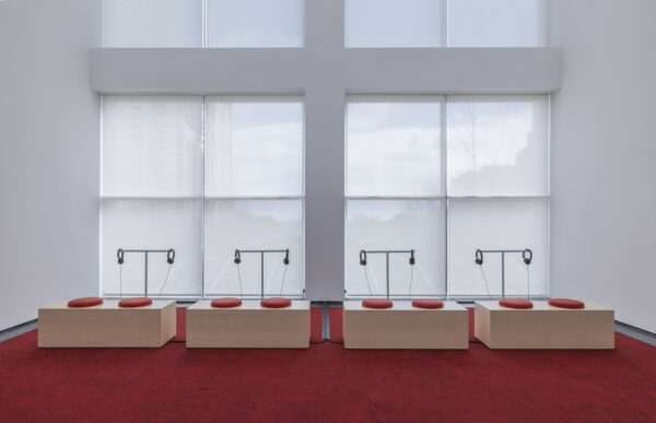 Four listening stations with two sets of head phones behind two cushions on wooden benches in a two story room filled with square windows and orange carpet
