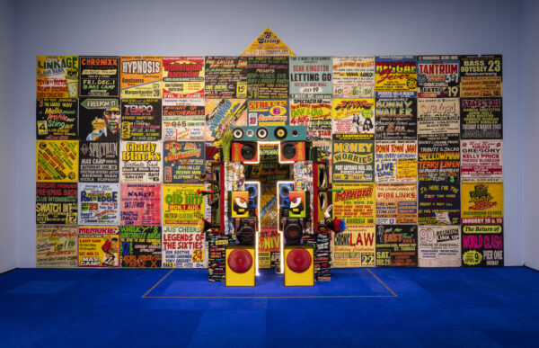Concert posters and speakers form a building shape in a blue room with bright blue carpet