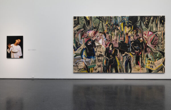 A large, expressive painting with dark, stylized figures amidst colorful, gestural brushstrokes hangs on a white museum wall. To its left, a smaller vertical photograph shows a person in a light garment with one hand raised, eyes closed, against a dark background.