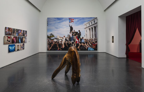 A white gallery space with a dynamic wooden sculpture in the foreground and photographs on the left and center walls. A red curtain frames a doorway to the right