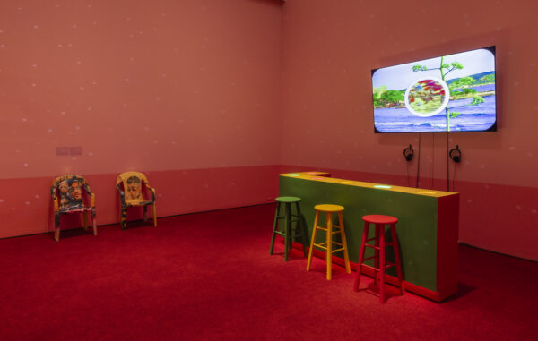 A colorful room with red carpeting contains a green and yellow bar-like counter with four multicolored stools. On the wall, a large screen displays a scene from the animated video. To the left, two lawn chairs feature images of Bad Bunny.