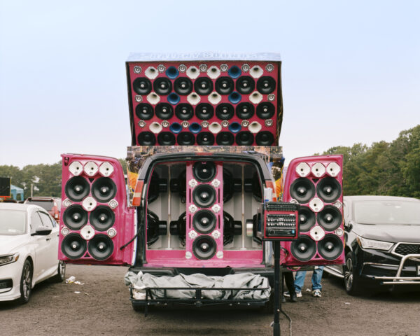 A van in a parking lot with its doors and roof opened up to reveal a hot pink interior and speakers covering every inch