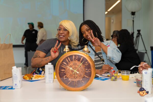Two people give peace signs to the camera from behind a giant pocket watch