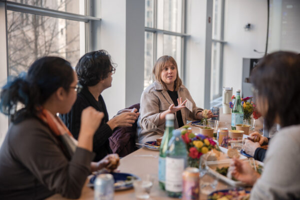 A group of people sit at a table filled with food and water in a room with floor to ceiling windows
