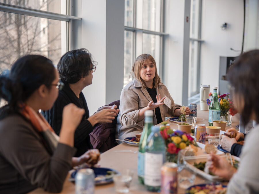 A group of people sit at a table filled with food and water in a room with floor to ceiling windows