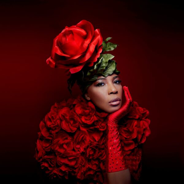 Against a deep garnet background a person with light-brown skin rests their chin on a gloved right hand, which complements a large rose headpiece and an outfit made of roses.