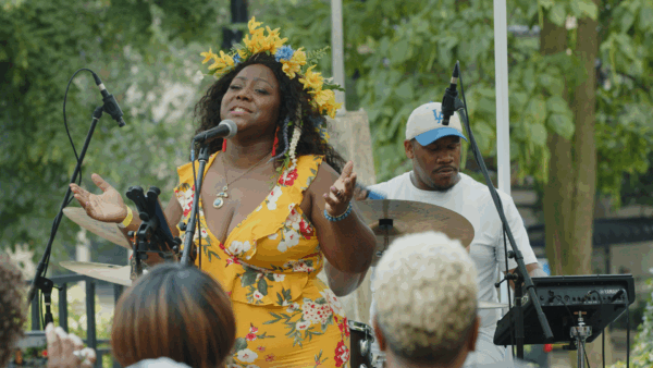 A Black woman in a floral yellow wrap dress and a wreath of yellow flowers on her head sings into a microphone with their eyes closed. A man in an LA cap sits behind her on the drums.