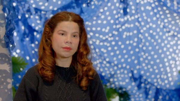A person with red curly hair sits in front of a blue painting with white polka dots