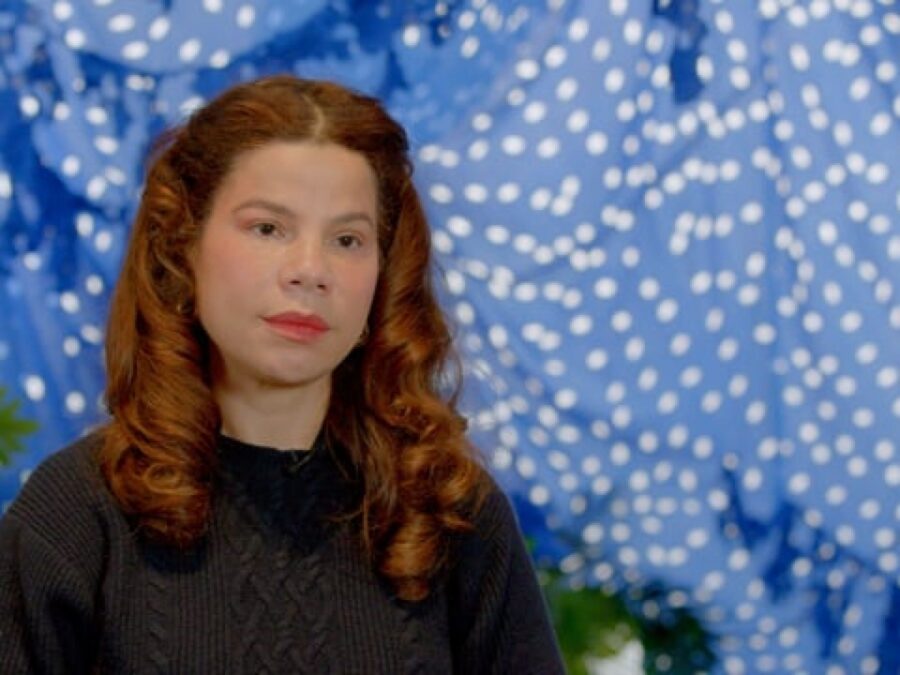 A person with red curly hair sits in front of a blue painting with white polka dots