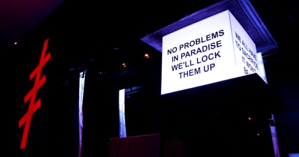 An illuminated white box at the top of the pillar with writing on two sides takes up a dark room with only a few lights in the background, including a red cross-hatch light. The closest side of the box reads No problems in paradise we'll lock them up.