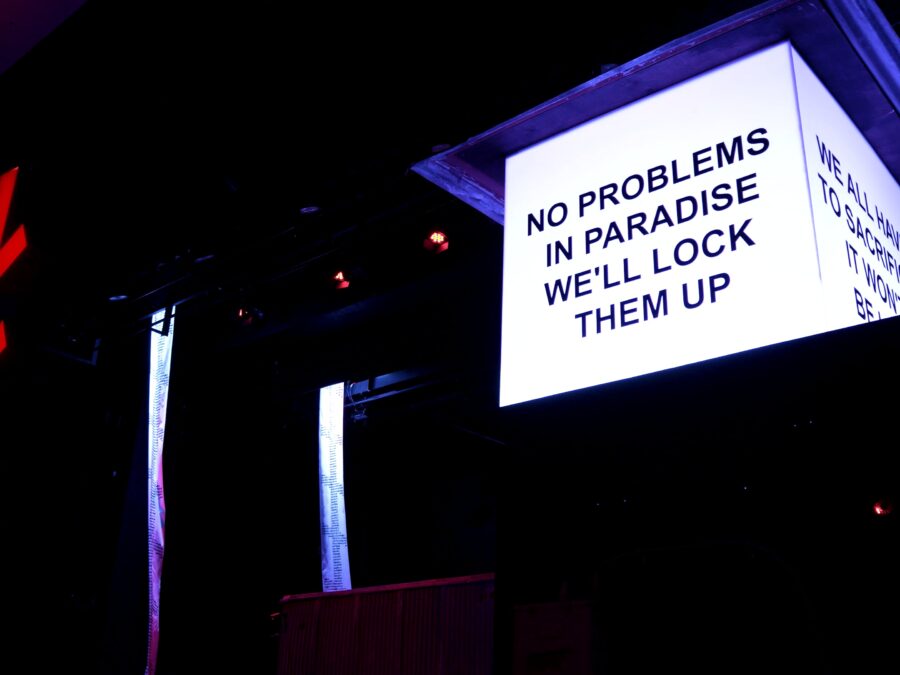 An illuminated white box at the top of the pillar with writing on two sides takes up a dark room with only a few lights in the background, including a red cross-hatch light. The closest side of the box reads No problems in paradise we'll lock them up.