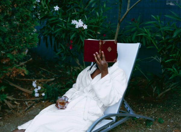 A person in a white robe lies on a white sunchair with a drink in their right hand resting on their leg. They hold a red book up with their left hand, obscuring their face. The backdrop is filled with tropical plants.