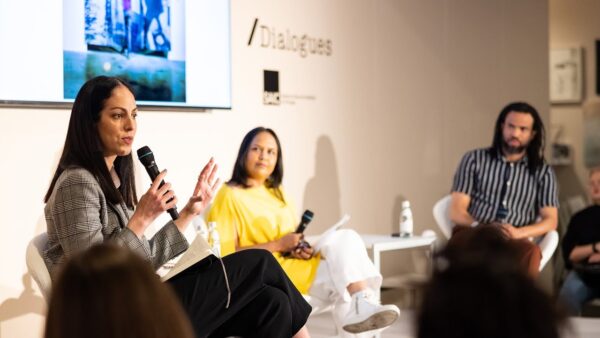 Three people sit in front of an audience in a cosy room with an artwork and the word Dialogues behind them