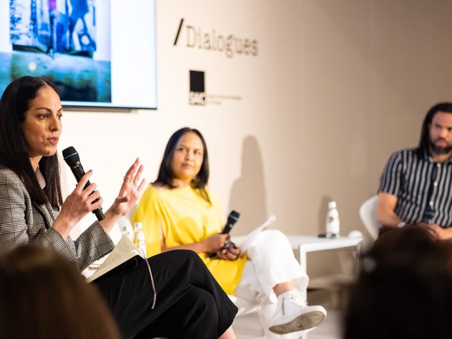 Three people sit in front of an audience in a cosy room with an artwork and the word Dialogues behind them