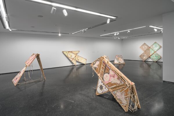A modern art gallery displays five geometric sculptures made of wood and fabric. The pieces are arranged throughout the room on a polished dark floor, with overhead lighting highlighting their intricate designs. The space is contemporary and minimalistic.