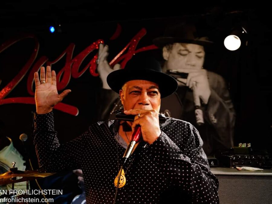 A male dressed in a black fedora and leather jacket holds a harmonica to their mouth in front of a mural of a musician holding the same pose