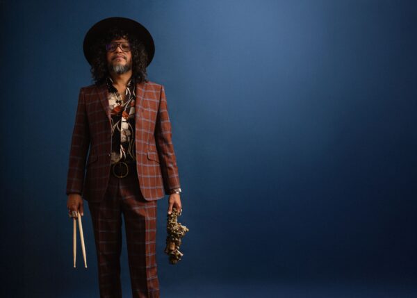 Moody photograph of a person in a checked suit and a hat holding drum sticks and a saxophone against a dark blue background