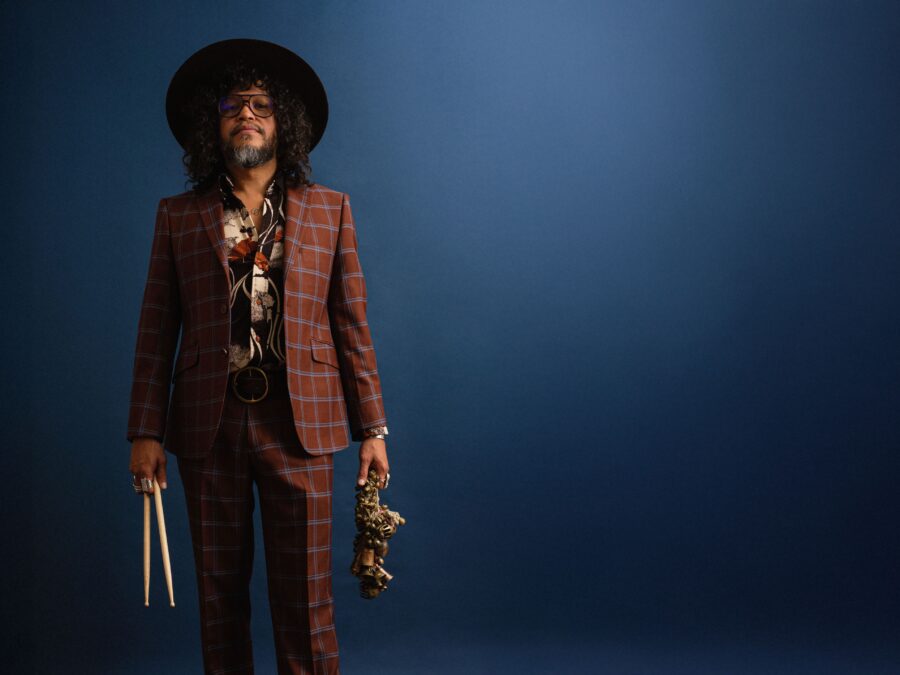 Moody photograph of a person in a checked suit and a hat holding drum sticks and a saxophone against a dark blue background