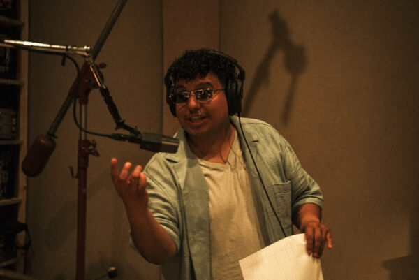 A person in a recording studio gestures with his right hand while in front of a microphone