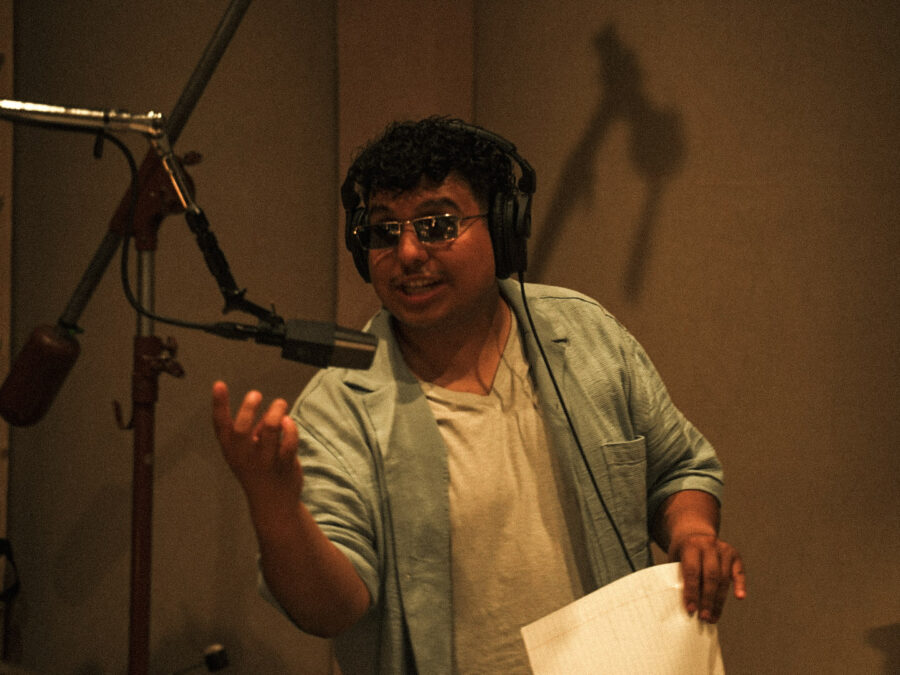 A person in a recording studio gestures with his right hand while in front of a microphone