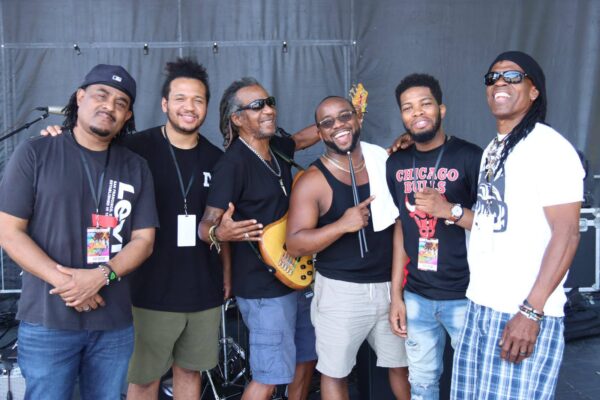 A group of people smile next to a grey wall, some of them carrying instruments