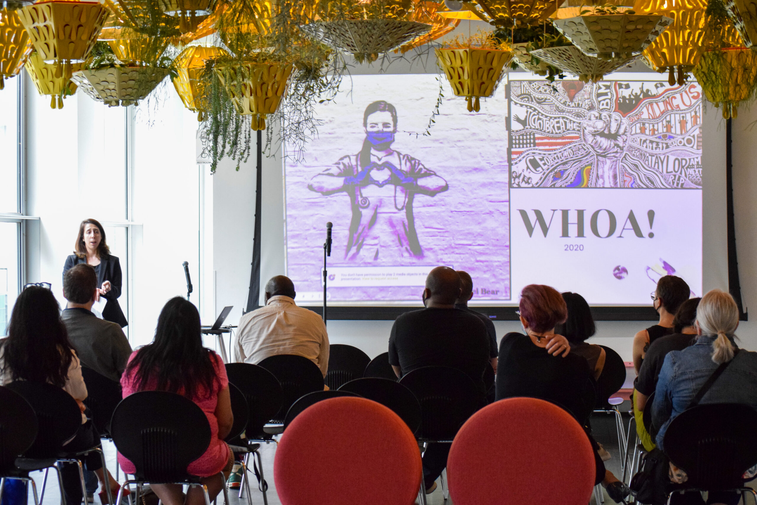 A group of people gather in front of a projection screen displaying a drawing of a masked medical professional making a heart with their hands in a room filled with hanging plants