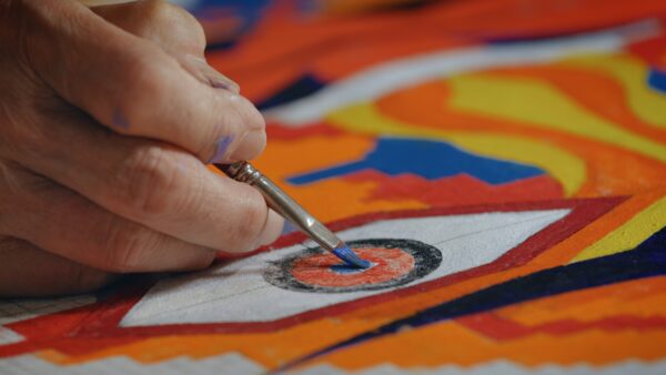 A hand is painting the pupil of an eye with a small brush within a colorful abstract design. The artwork features vibrant colors like orange, red, blue, and yellow in dynamic shapes and lines.