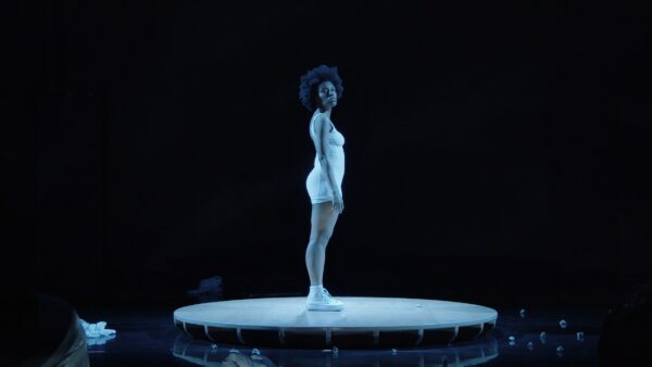 A performer stands on a circular stage in a dimly lit setting, with a spotlight creating a dramatic effect. The figure stands erect their body facing stage left, but their head turned forward. The background is dark, highlighting the stage as the focal point.
