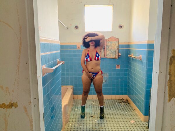 A person wearing a Puerto Rican flag bikini poses in a blue tiled bathroom with their hands covering their eyes and their pubic region